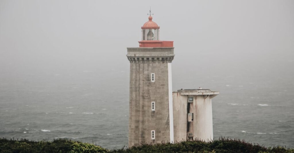 Lighthouse stands tall amidst foggy and overcast conditions by the sea, offering navigational guidance.